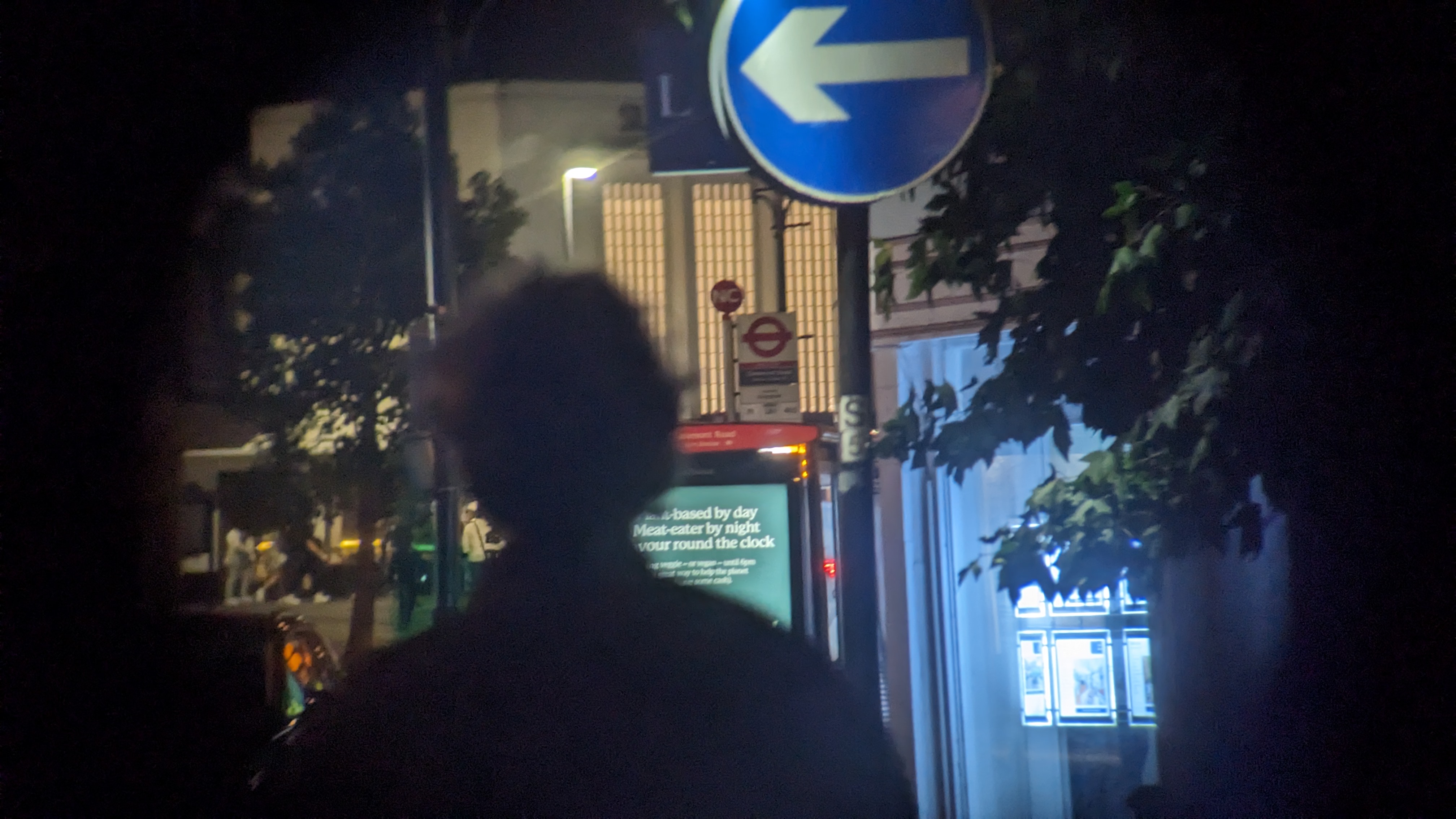 A photograph taken through binoculars of a silhouetted figure walking down a high street at night with traffic signs, a bus stop and trees around them.
